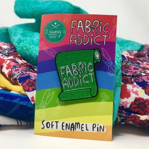 A green sparkly enamel pin badge in the shape of a roll of fabric, with the words Fabric Addict in white.