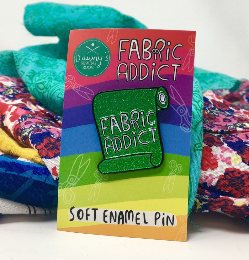 A green sparkly enamel pin badge in the shape of a roll of fabric, with the words Fabric Addict in white.