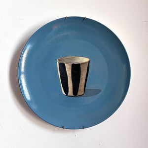 May include: A painting of a blue plate with a black and white striped cup. The plate is a solid, matte blue, and the cup has vertical black stripes. The cup casts a shadow on the plate.