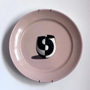 May include: A decorative plate with a pale pink surface. The plate features a black and white abstract design of a vase or cup. The plate is mounted on a white wall with three small metal hooks.
