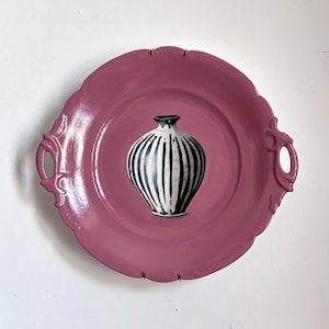 May include: A decorative plate in a muted rose colour with a scalloped edge and ornate handles. A small vase with black and white vertical stripes is centered on the plate. The vase has a black rim.