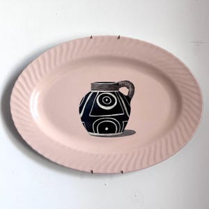 May include: A pink oval serving platter with a textured edge, displaying a black and white geometric design jug with a woven handle. The jug features circles and curved lines. The platter is set against a white backdrop.