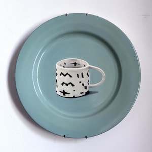 May include: A white ceramic mug with black abstract shapes sits on a large, round, pale blue plate. The mug has a handle and is decorated with various geometric patterns. The plate is mounted on a white wall.