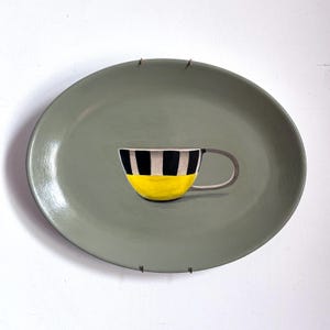 May include: A painted ceramic plate with a cup design. The cup is yellow with black and white stripes and a grey handle. The plate is oval and painted in a muted green colour.