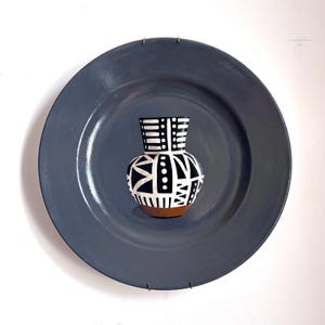 May include: A small, decorative vase with a black and white geometric pattern, set on a large, grey plate. The vase has a brown base and is centered on the plate, which is mounted on a white wall.