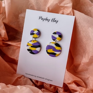 May include: Purple, yellow, black, and white patterned clay earrings. The earrings are double-layered and have a round shape. The card reads "Payday Clay" and "handmade by elizabeth dingman".