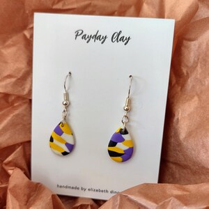 May include: A pair of dangle earrings with a teardrop-shaped purple, yellow, black, and white patterned design. The earrings are on a white card with the text "Payday Clay" and "handmade by elizabeth ding".