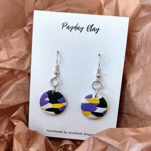 May include: A pair of handmade earrings with a circular design. The earrings feature a pattern of purple, yellow, black, and white shapes. The text "Payday Clay" is at the top of the card, and "handmade by elizabeth dingm" is at the bottom.