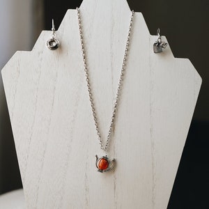 May include: A silver necklace with a teapot pendant featuring a red gemstone. The necklace is displayed on a white jewelry stand with a pair of silver earrings.