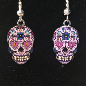 May include: A pair of silver dangle earrings with pink sugar skull charms. The skulls have black outlines and colorful floral details. The eyes are pink hearts with black outlines.