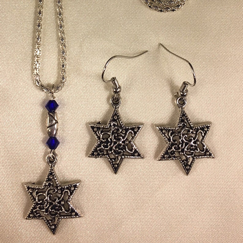 May include: A silver necklace with a blue crystal charm and a silver pendant in the shape of a Star of David. Matching silver earrings with Star of David pendants.