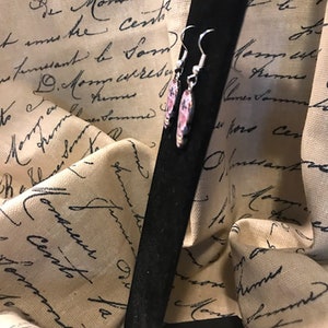 May include: A pair of silver earrings with pink and white floral designs. The earrings are hanging from a black background.