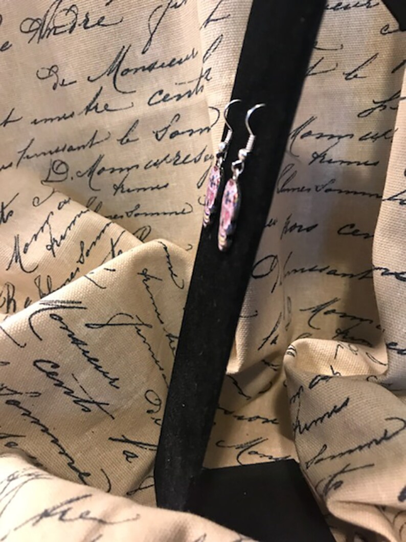 May include: A pair of silver earrings with pink and white floral designs. The earrings are hanging from a black background.