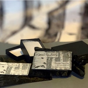 May include: Two black boxes with a white and black patterned business card that says "Aspen Tree Jewelry" and "Francie Ann Cole". The boxes are on a gray surface with a black velvet bag.