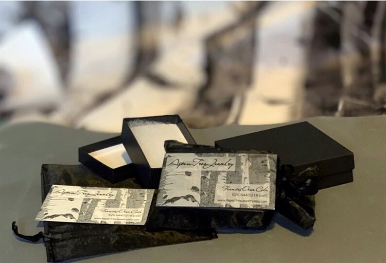 May include: Two black boxes with a white and black patterned business card that says "Aspen Tree Jewelry" and "Francie Ann Cole". The boxes are on a gray surface with a black velvet bag.