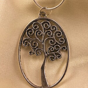 May include: A silver necklace with a pendant shaped like a tree with a swirling design. The pendant is oval shaped and hangs from a silver chain.