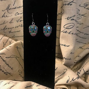 May include: A pair of silver dangle earrings with a sugar skull design. The skulls are green, blue, pink, and black with floral patterns.