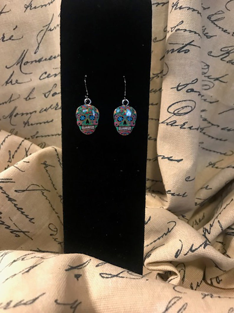 May include: A pair of silver dangle earrings with a sugar skull design. The skulls are green, blue, pink, and black with floral patterns.