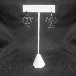 May include: A pair of silver menorah earrings hanging on a white display stand against a black background.