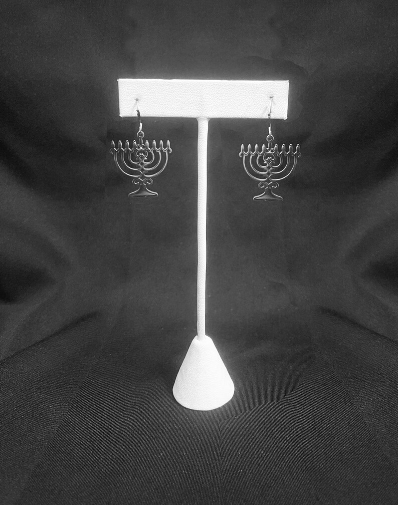 May include: A pair of silver menorah earrings hanging on a white display stand against a black background.