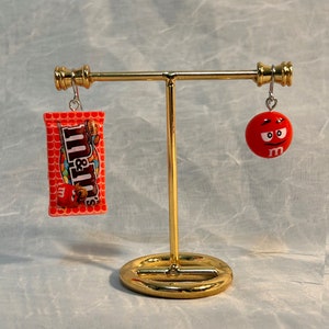 May include: A pair of earrings on a gold stand. The left earring is a red and white M&M's candy bar. The right earring is a red M&M's candy with a white M and a face.