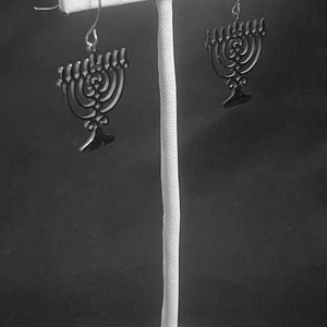 May include: A pair of silver menorah earrings on a white display stand. The menorahs are black and have a detailed design.