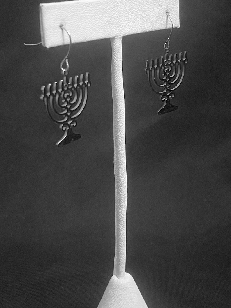May include: A pair of silver menorah earrings on a white display stand. The menorahs are black and have a detailed design.