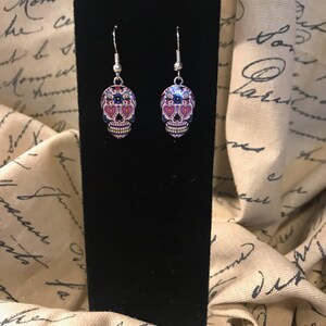May include: A pair of silver dangle earrings with pink, blue, and black sugar skull charms.