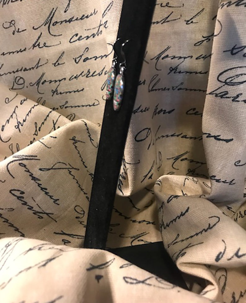 May include: A pair of dangle earrings with a colorful, speckled design. The earrings are hanging from a black stand against a background of fabric with black script.