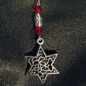May include: A silver charm in the shape of a Star of David with intricate filigree details. The charm is attached to a silver chain with red crystal beads.