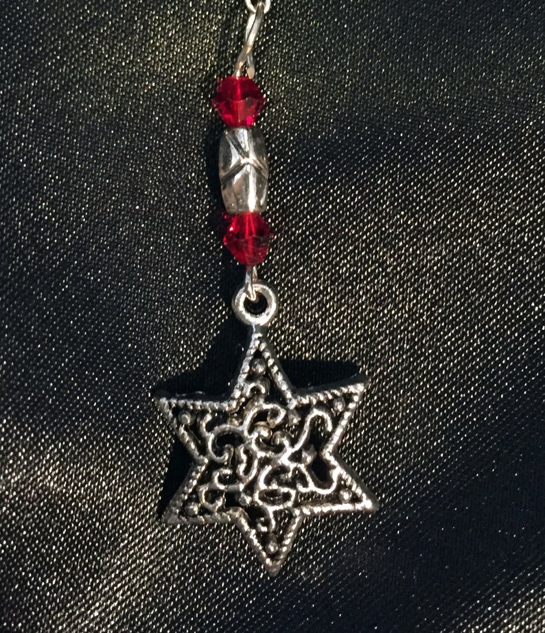 May include: A silver charm in the shape of a Star of David with intricate filigree details. The charm is attached to a silver chain with red crystal beads.