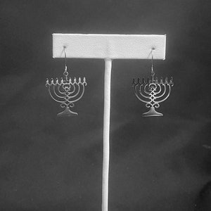 May include: A pair of silver menorah earrings hanging on a white display stand against a black background.