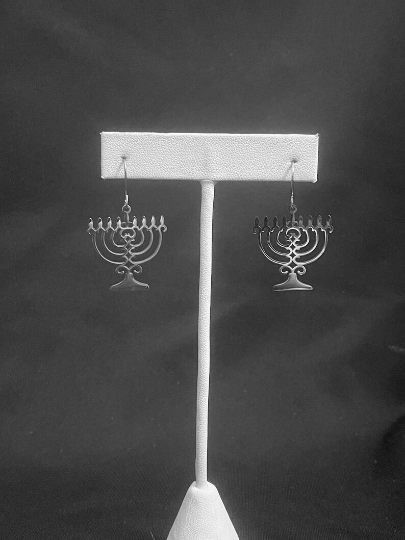 May include: A pair of silver menorah earrings hanging on a white display stand against a black background.