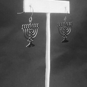 May include: A pair of silver menorah earrings hanging on a white display stand. The earrings are detailed with a delicate, intricate design.
