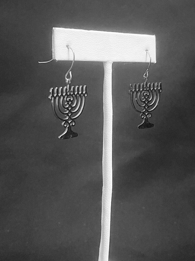 May include: A pair of silver menorah earrings hanging on a white display stand. The earrings are detailed with a delicate, intricate design.