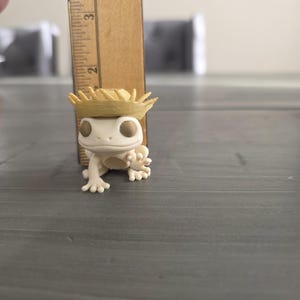 May include: A small, off-white gecko figurine wearing a miniature straw hat. The gecko is positioned on a gray surface, with a wooden ruler next to it. The ruler measures the gecko's height, approximately 2 inches.