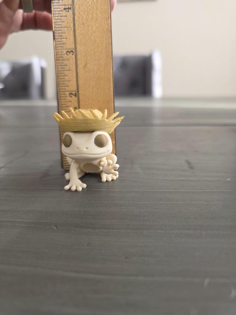 May include: A small, off-white gecko figurine wearing a miniature straw hat. The gecko is positioned on a gray surface, with a wooden ruler next to it. The ruler measures the gecko's height, approximately 2 inches.