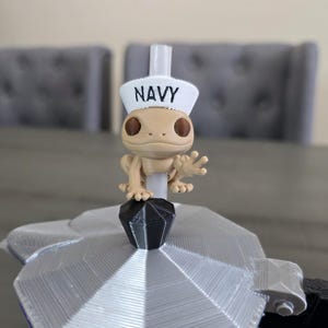 May include: A tan figurine of a gecko wearing a white sailor hat with the word "NAVY" printed on it. The gecko is perched on a silver and black object, possibly a coffee maker. The background includes two gray chairs.