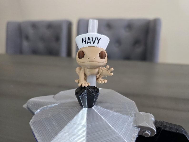 May include: A tan figurine of a gecko wearing a white sailor hat with the word "NAVY" printed on it. The gecko is perched on a silver and black object, possibly a coffee maker. The background includes two gray chairs.