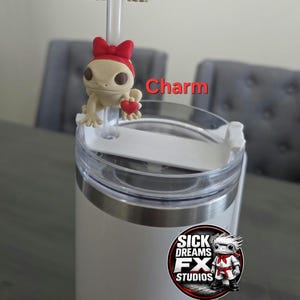 May include: A white tumbler with a clear lid and a charm. The charm is a beige gecko wearing a red bow and holding a red heart. The text "Charm" is visible. The tumbler has a logo that says "Sick Dreams FX Studios".