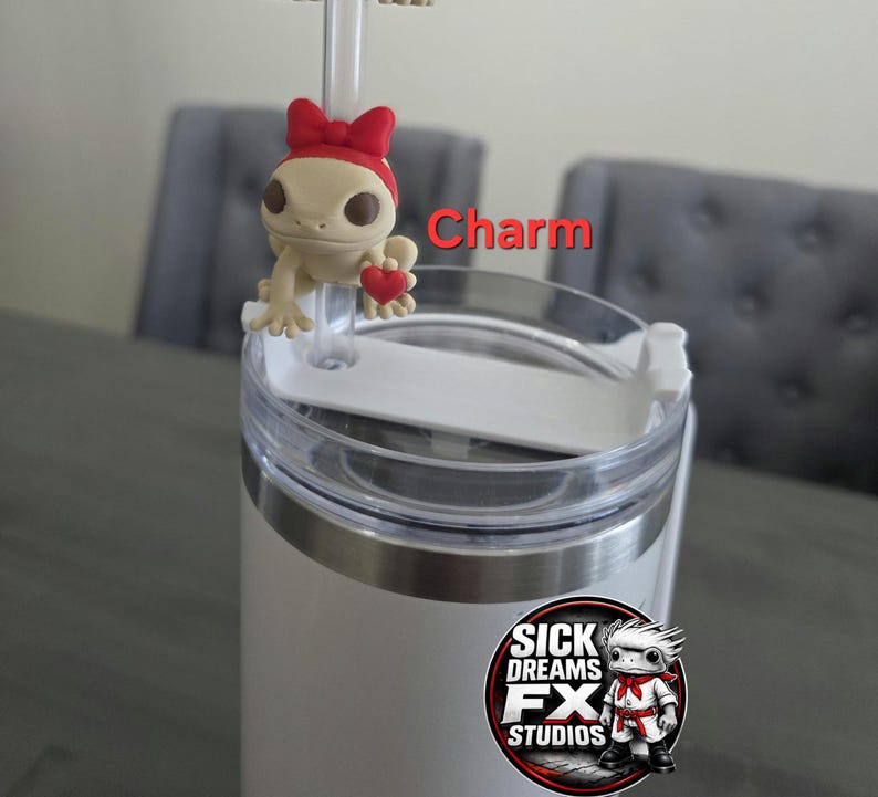 May include: A white tumbler with a clear lid and a charm. The charm is a beige gecko wearing a red bow and holding a red heart. The text "Charm" is visible. The tumbler has a logo that says "Sick Dreams FX Studios".