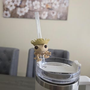 May include: A white and silver Stanley tumbler with a clear straw and a small, tan frog figurine attached. The frog is wearing a straw hat. The tumbler is on a gray table.