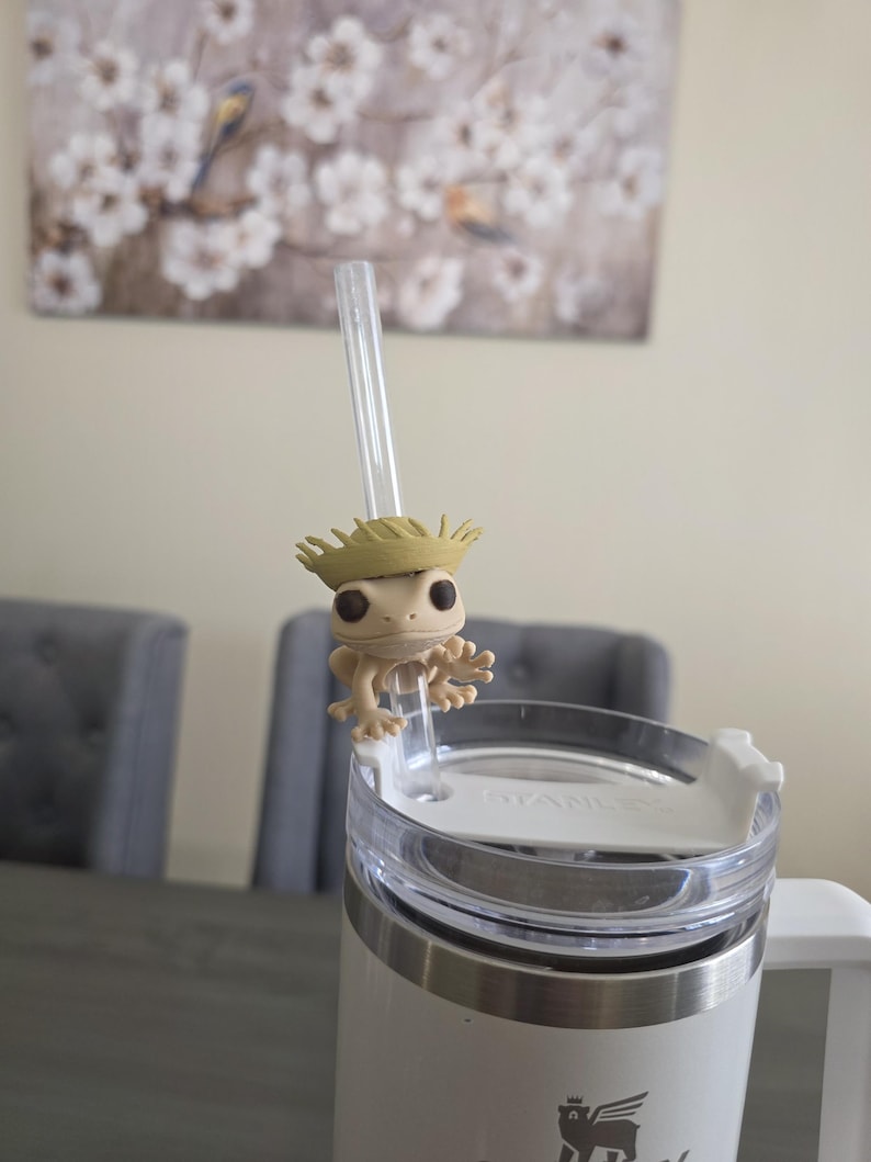 May include: A white and silver Stanley tumbler with a clear straw and a small, tan frog figurine attached. The frog is wearing a straw hat. The tumbler is on a gray table.
