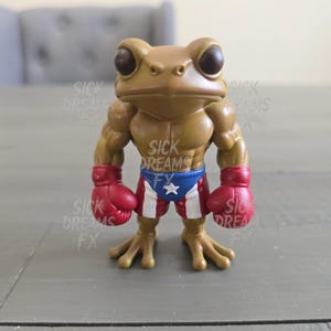 May include: A tan frog figurine with muscular arms, wearing red boxing gloves and Puerto Rican flag-themed shorts. The figurine has large, dark eyes and is posed in a standing position. The words "SICK DREAMS FX" are visible.