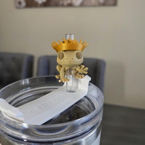 May include: A tan gecko figurine wearing a gold crown sits atop a white lid of a stainless steel tumbler. The tumbler has the word "STANLEY" printed on the lid. The background includes a painting and two gray chairs.