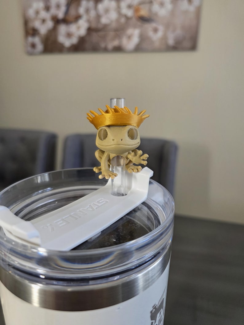 May include: A tan gecko figurine wearing a gold crown sits atop a white lid of a stainless steel tumbler. The tumbler has the word "STANLEY" printed on the lid. The background includes a painting and two gray chairs.