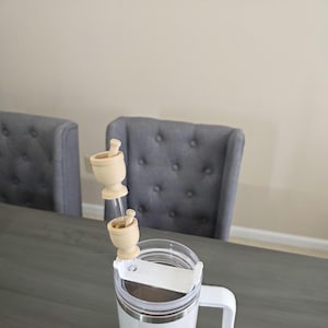 May include: A white Stanley mug with a clear lid and a white handle. Two small wooden mortar and pestle sets are attached to the lid. The background includes a gray table and two gray upholstered chairs.