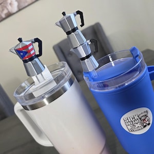 May include: Two insulated coffee mugs. One is white with a silver rim and handle, topped with a miniature Puerto Rican flag-themed coffee maker. The other is blue with a handle and a sticker that says "Sick Dreams FX Studios", topped with a miniature coffee maker.