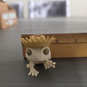 May include: A small, off-white figurine of a frog wearing a tan straw hat. The frog has large, dark eyes and outstretched arms. A wooden ruler is in the background.