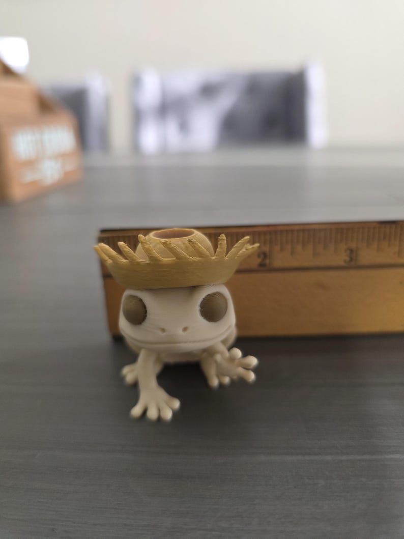 May include: A small, off-white figurine of a frog wearing a tan straw hat. The frog has large, dark eyes and outstretched arms. A wooden ruler is in the background.